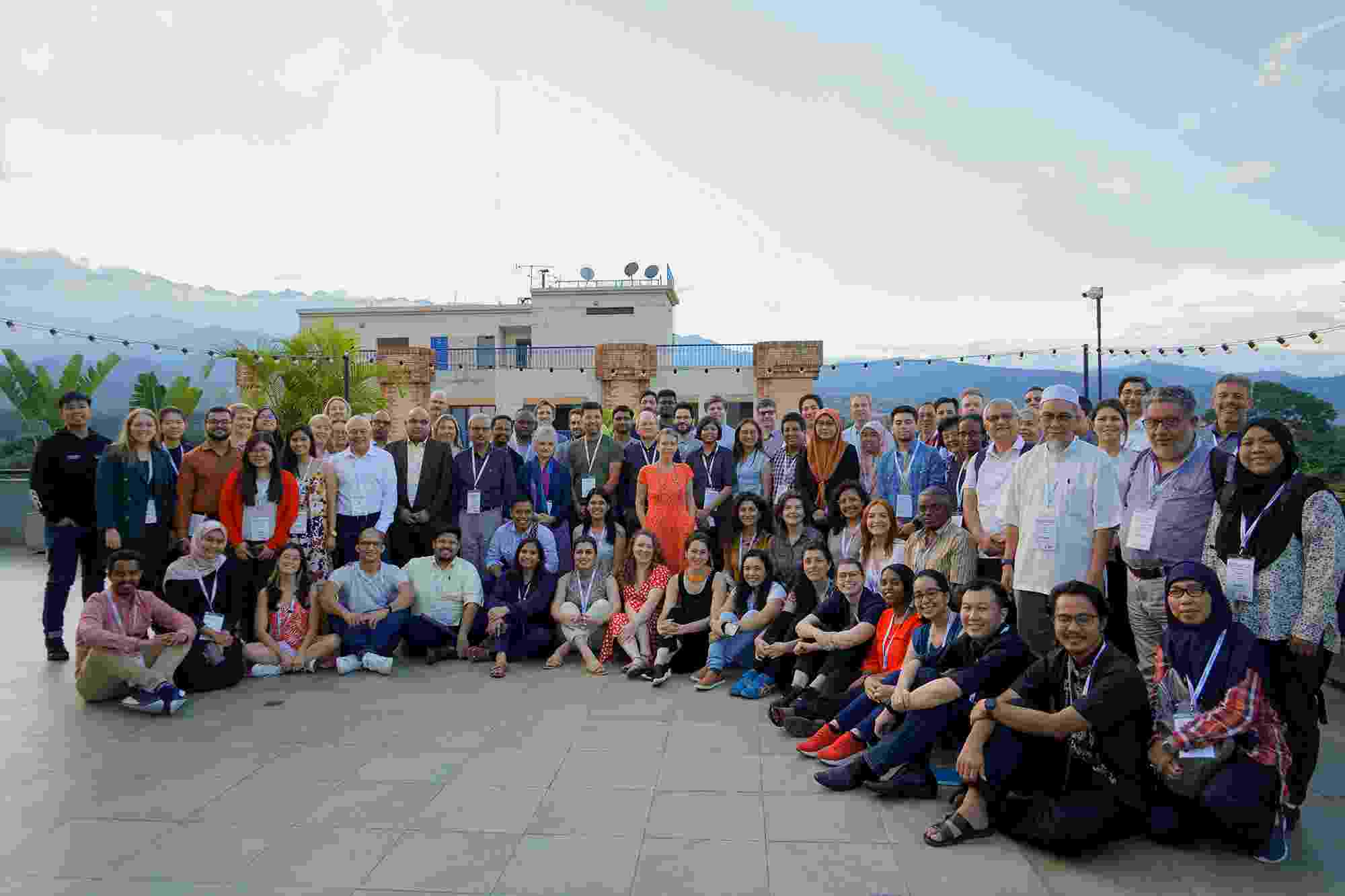 A large mixed group of Hub members sit and stand together in several rows, smiling at the camera, with rooftops and blue sky visible behind them